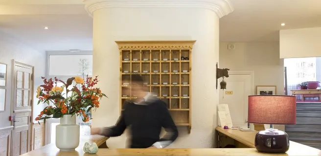 Réception d'hôtel lumineuse et accueillante. Derrière un grand comptoir en bois clair, on peut apercevoir une personne souriante. Des fleurs colorées dans un vase apportent une touche de fraîcheur à l'espace.
